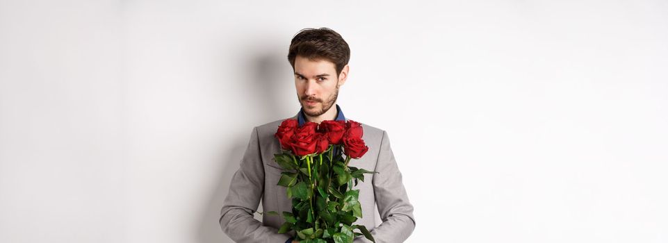 Handsome macho man going on date in suit, holding bouquet of red roses and smiling at camera, making valentines day surprise gift, white background.