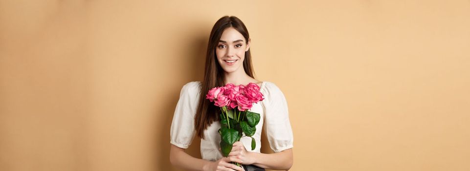 Romantic Valentines day concept. Beautiful young lady holding pink roses and smiling, standing happy on beige background.