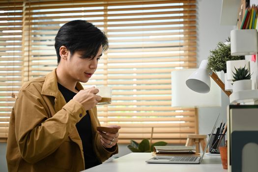 Young Asian man drinking coffee and reading email on laptop computer.