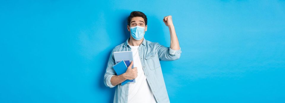 Education, covid-19 and social distancing. Excited male student in medical mask triumphing, raising fist up and holding notebooks, standing over blue background.
