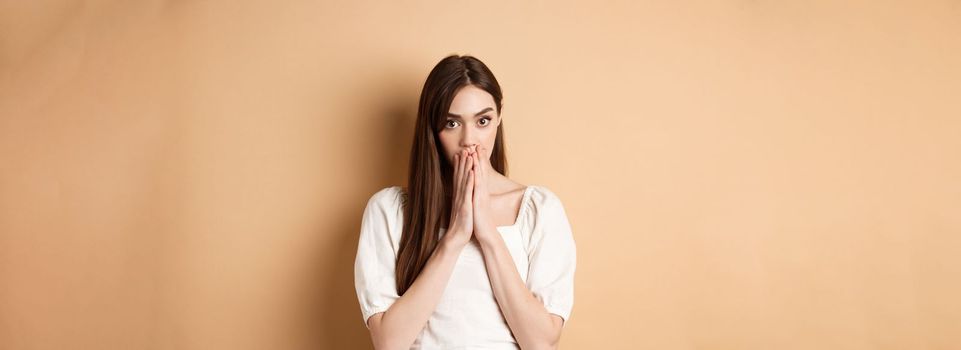 Omg shocking. Speechless woman covering mouth with hands and staring shocked at camera, gossiping, hear stunning news, beige background.