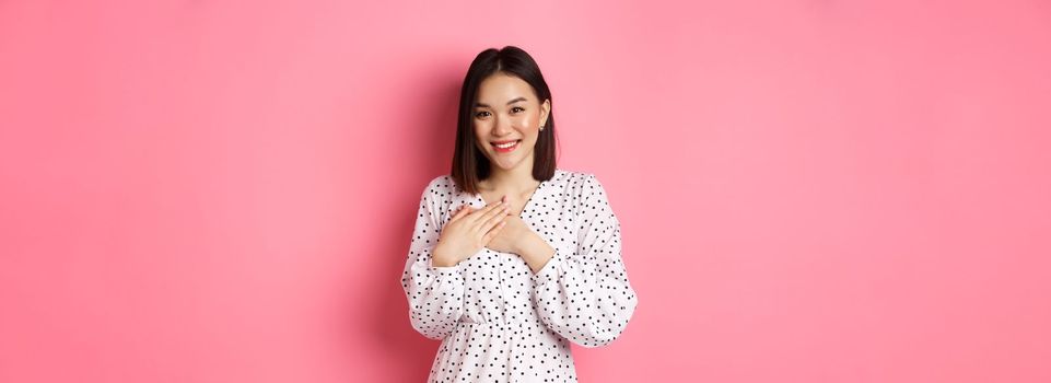 Thankful Korean girl in dress smiling, holding hands on heart and looking grateful at camera, touched with nice gesture, standing over pink background.