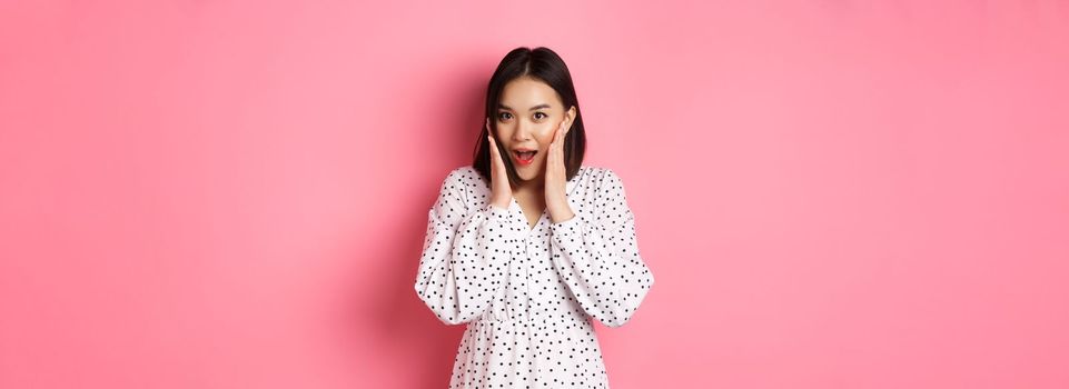 Young asian woman looking intrigued, gossiping and saying wow with curious face, looking interested at camera, standing over pink background.