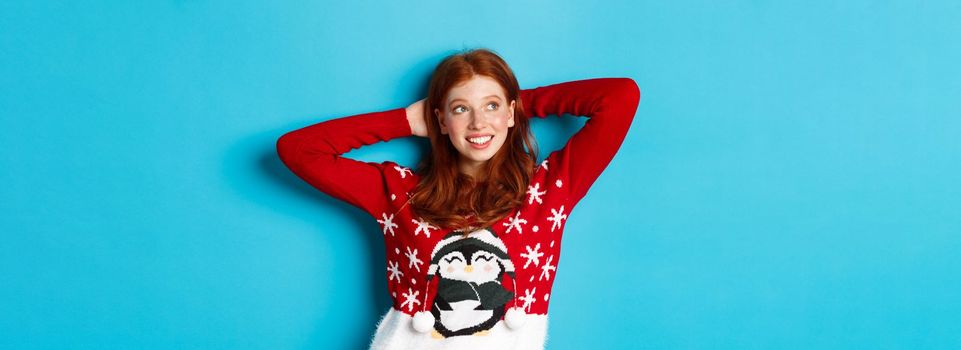 Winter holidays and celebration concept. Relaxed teenage girl holding hands behind head and looking left at promo, resting on Christmas Eve, standing over blue background.