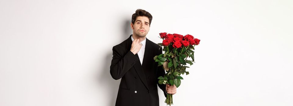 Image of elegant and sassy man in black suit, looking confident and holding bouquet of red roses, going on a romantic date, standing against white background.