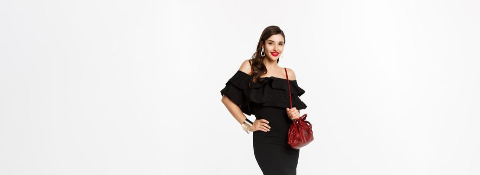 Beauty and fashion concept. Full length of elegant young woman going shopping in black dress, heels and purse, looking confident, standing over white background.