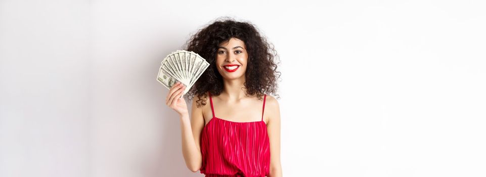 Stylish smiling woman in red dress showing money, standing on white background.