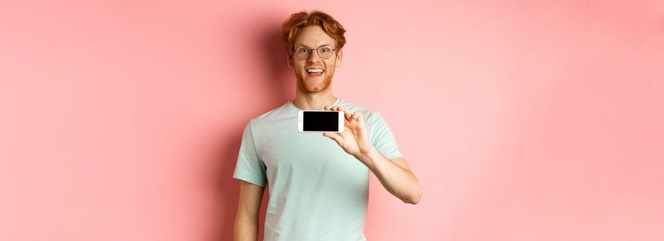 Happy young man with red hair showing smartphone screen, holding phone horizontally and smiling amazed, standing over pink background.