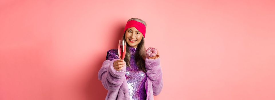 Celebration and holidays concept. Stylish asian elderly woman having fun on party, eating sweet donut and extending hand with glass of champagne, saying cheers, pink background.