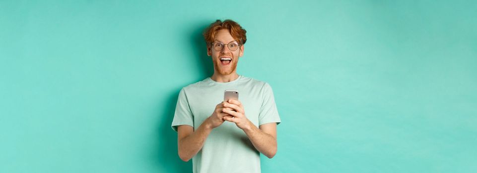 Surprised redhead man in glasses looking amazed at camera after reading promo offer on smartphone, standing against turquoise background.