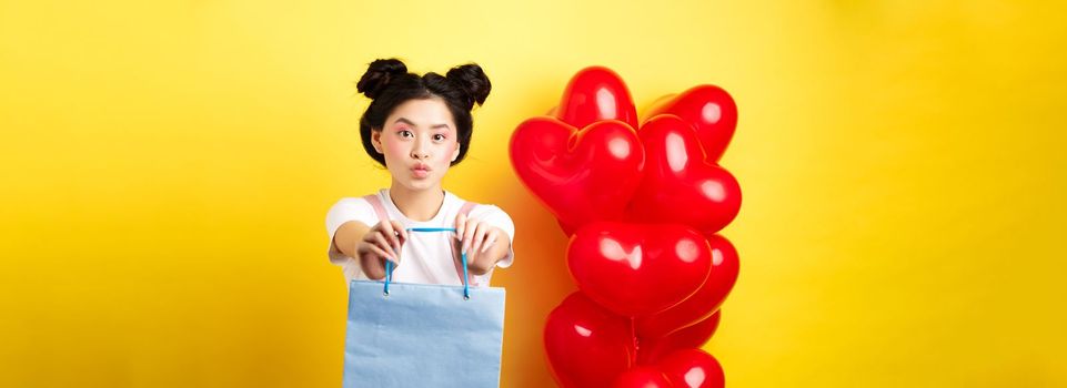 Happy Valentines day. Cute asian girlfriend giving lover a gift, stretch out hands with shopping bag and looking silly, standing over yellow background near heart balloons.