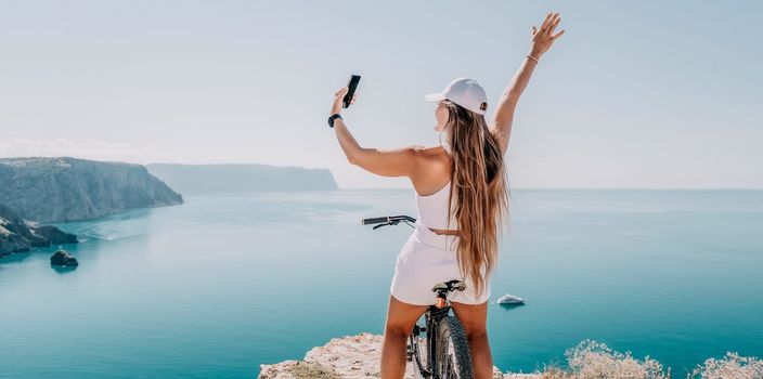 A woman cyclist on a mountain bike looking at the landscape of mountains and sea. Rear view of cyclist woman standing in front to the sea with outstretched arms. Freedom and healthy lifestyle concept