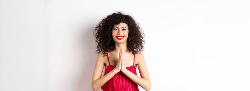 Attractive young woman with red lips, evening dress, saying thank you and smiling, looking grateful with namaste gesture, standing over white background.