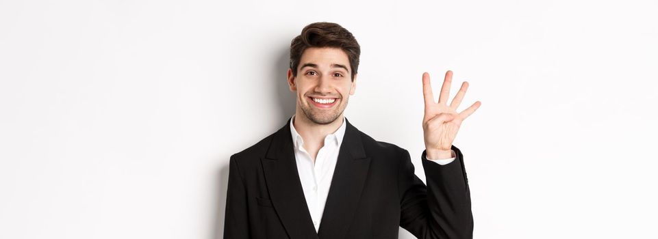 Close-up of handsome businessman in black suit, smiling amazed, showing number four, standing over white background.