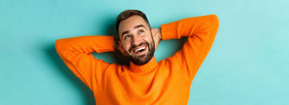 Carefree young man daydreaming, resting with hands behind head and looking at upper left corner happy, standing over turquoise background in orange sweater.