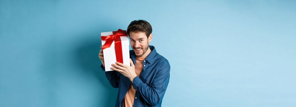 Valentines day. Romantic boyfriend showing gift box for lover, celebrating anniversary, standing over blue background.