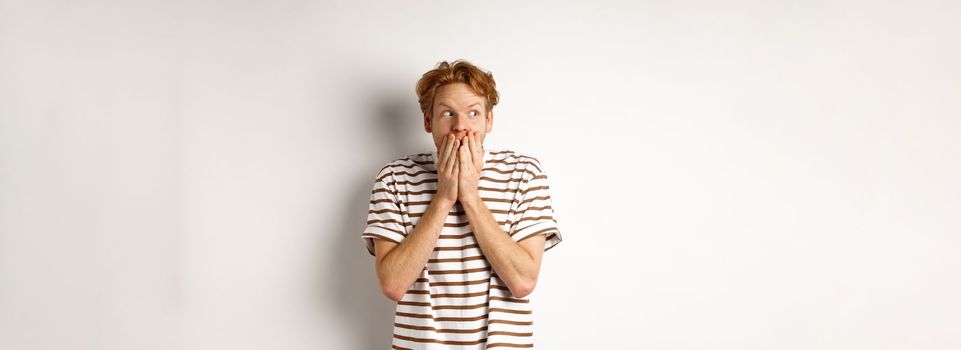 Shocked redhead guy gossiping, giggle in hands and looking left impressed, standing over white background.