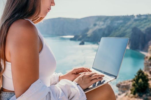 Digital nomad, Business woman working on laptop by the sea. Pretty lady typing on computer by the sea at sunset, makes a business transaction online from a distance. Freelance remote work on vacation