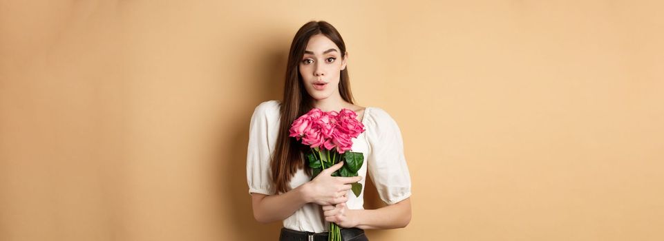 Valentines day. Image of surprised girlfriend thanking for flowers, receive pink roses from lover and looking grateful at lover, standing on beige background.