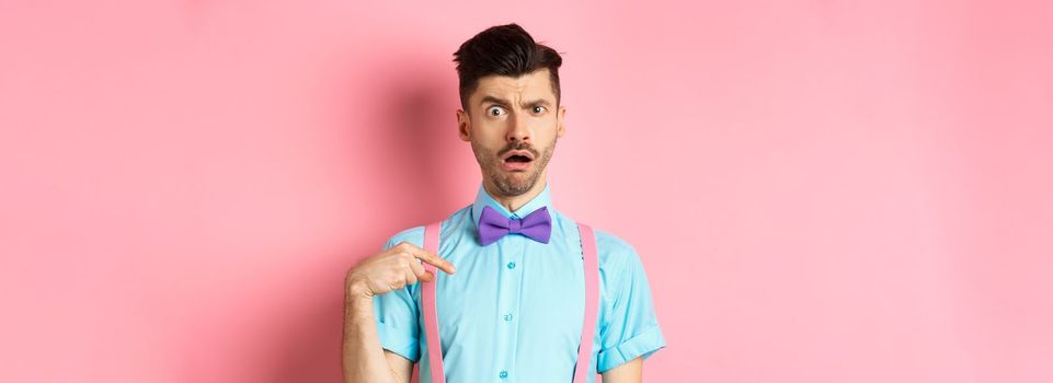 Image of confused funny guy in bow-tie and suspenders, pointing at himself as if being accused or chosen, raising eyebrow surprised, standing over pink background.
