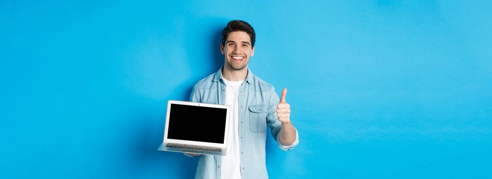 Smiling handsome man showing laptop screen and thumb-up, like promo offer, recommending website, standing against blue background.