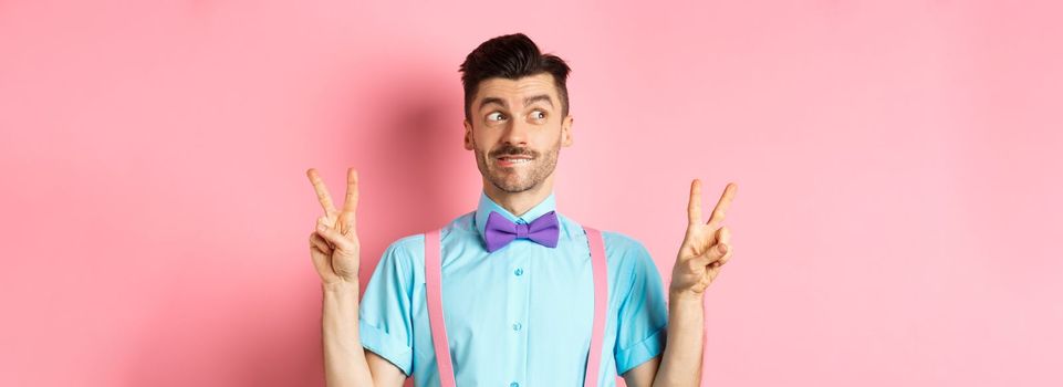 Funny young guy in bow-tie, entertain people on holiday event, showing peace signs and looking aside with excitement, standing over pink background.