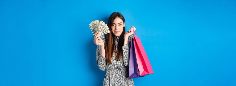 Shopping. Excited attractive woman buying gifts for herself, showing dollar bills and color bags from shop, smiling satisfied, standing on blue background.