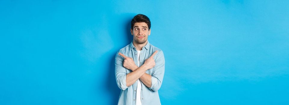 Portrait of indecisive adult man pointing fingers sideways but looking left, making choice between two products, standing against blue background.