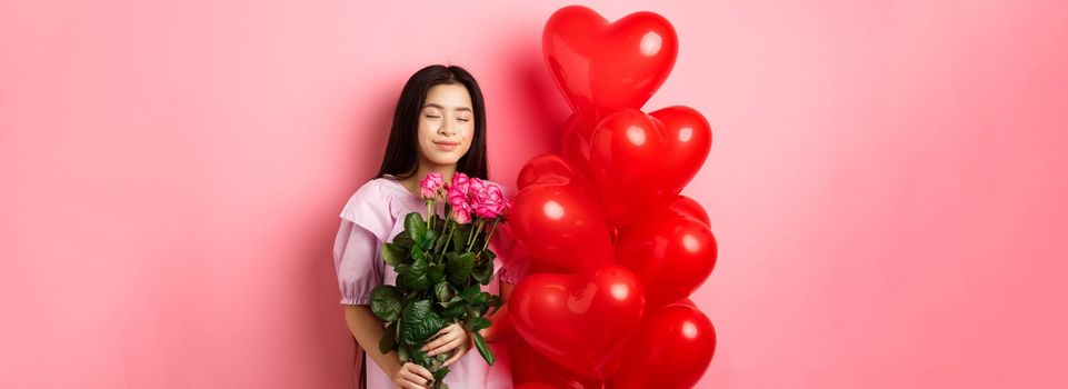 Valentines day concept. Romantic teen asian girl dreaming of love or date, close eyes and smile, holding flowers from lover, receive bouquet of roses and red heart balloons, pink background.
