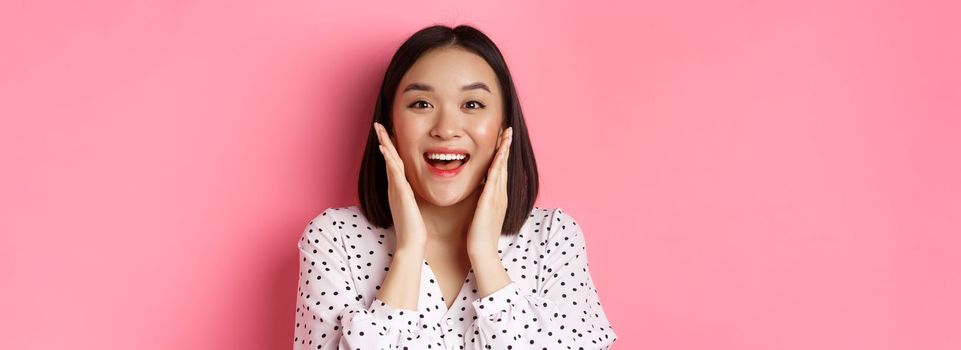 Close-up of beautiful asian woman touching clean skin face and staring at camera amazed, standing over pink background.