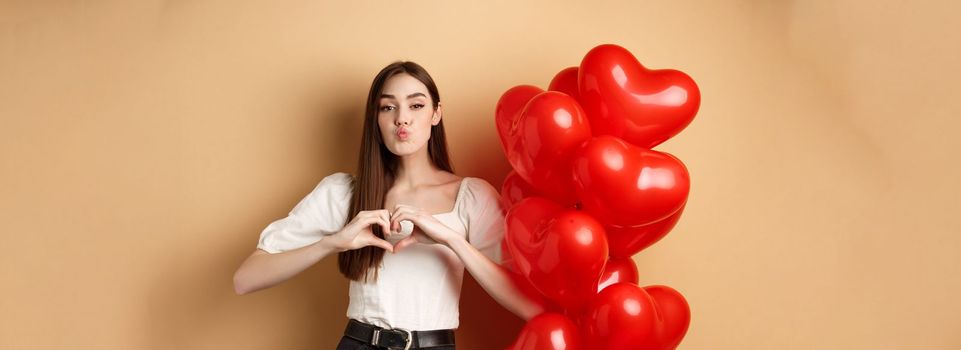 Lovely girlfriend pucker lips and waiting for romantic kiss, showing heart gesture to say I love you, look at lover, standing near Valentines day balloons and beige background.