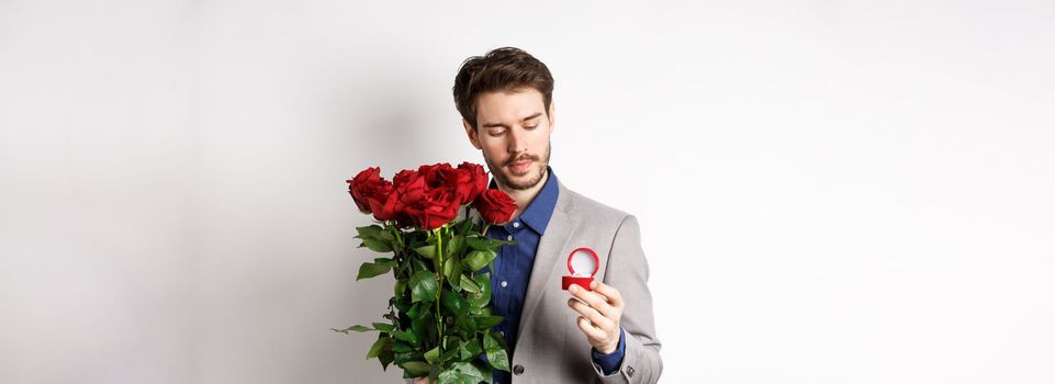 Romantic man in suit looking pensive at engagement ring, going to make a marriage proposal on Valentines day, holding bouquet of roses, standing over white background.