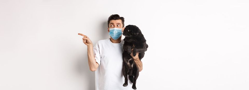 Covid-19, animals and quarantine concept. Young man in face mask holding cute black pug, dog and pet owner looking right at copy space, standing over white background.