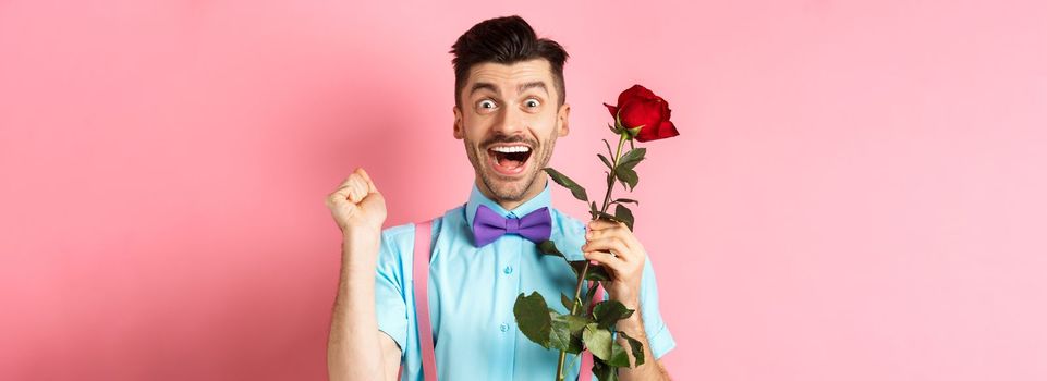 Valentines day and romance concept. Excited boyfriend jumping from happiness on romantic date, holding red rose and celebrating, standing on pink background.