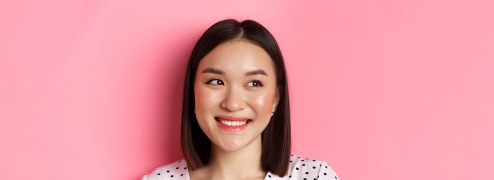 Beauty and lifestyle concept. Headshot of pretty asian woman dreamy gazing left at copy space, smiling happy, standing over pink background.