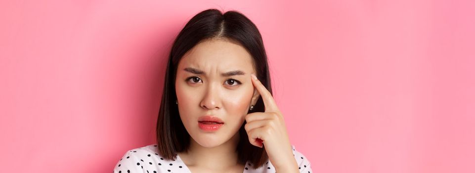 Beauty and skin care concept. Headshot of cute asian woman staring confused, pointing finger at head at frowning, standing over pink background.