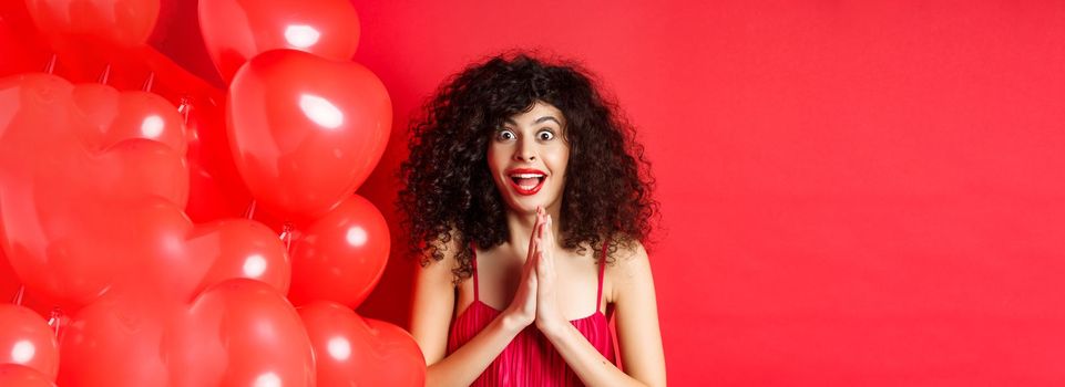 Valentines day. Excited caucasian woman in dress jumping from amazement, looking at something taunting, want to get product, standing near hearts balloons, white background.