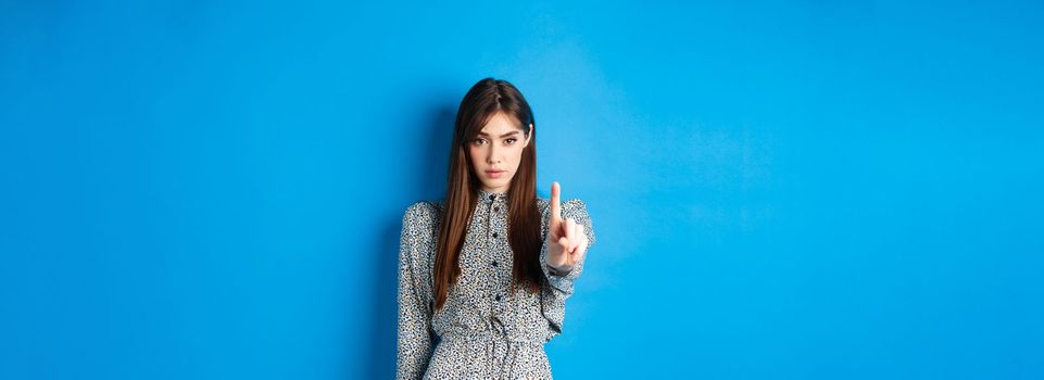 Serious woman looking confident and shaking finger, say no, stop you, disagree and disapprove, standing in dress on blue background.