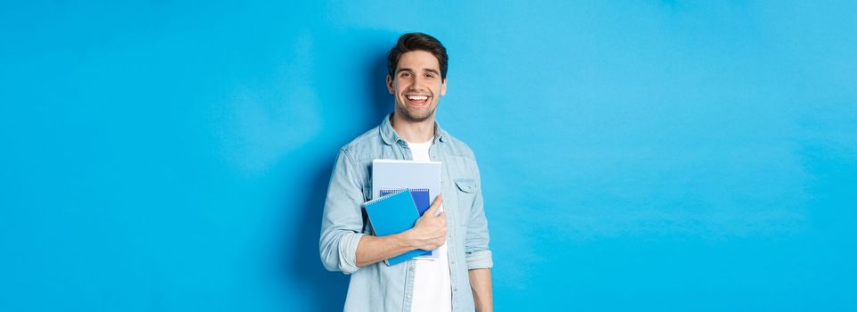 Smiling man studying, holding notebooks and looking happy, standing over blue background.