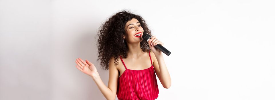 Elegant curly-haired woman in red dress singing in microphone, looking aside and smiling happy, standing on white background.