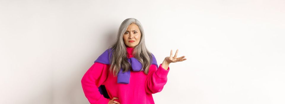 Angry and confused asian senior woman spread hand sideways and staring at camera puzzled, standing in pink sweater against white background.