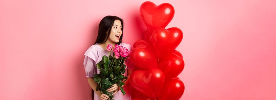 Portrait of asian teenage girl in love, holding flowers and looking at valentines day heart balloons, being on romantic date, pink background.