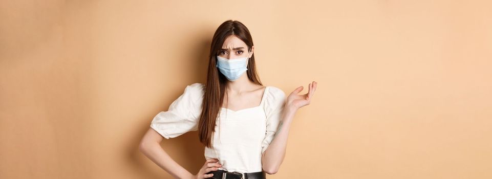 Covid-19 and lifestyle concept. Confused girl in medical mask frowning, pointing left with complaint, disappointed with something bad, standing against beige background.