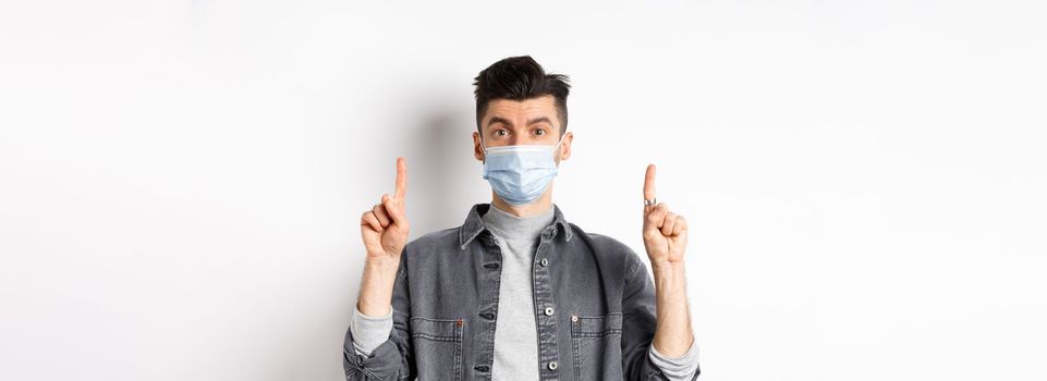 Pandemic lifestyle, healthcare and medicine concept. Stylish modern man in medical mask showing advertisement, pointing fingers up at logo, standing on white background.
