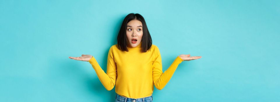 Confused and shocked asian woman dont understand, shrugging with spread hands, gasping and looking left, know nothing, standing over blue background.