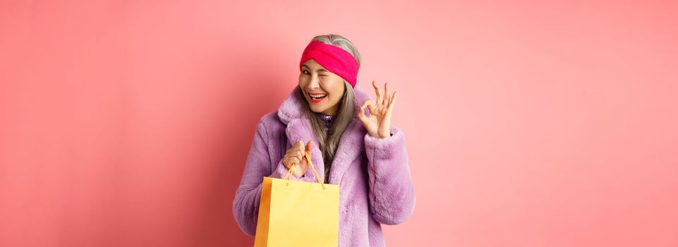 Stylish senior asian woman recommending store, holding shopping bag and winking with OK sign, smiling satisfied, standing over pink background.