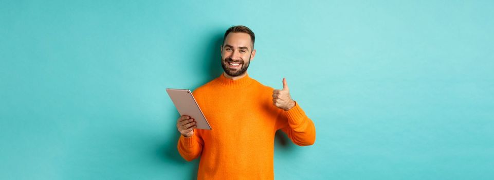 Satisfied adult man smiling, using digital tablet and showing thumb-up, approve and agree, standing against turquoise background.