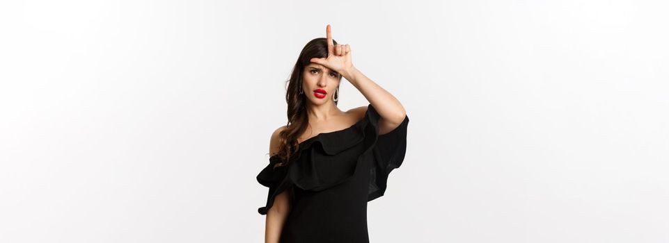 Fashion and beauty. Arrogant glamour woman showing loser sign on forehead, mocking lost person, standing in black dress over white background.