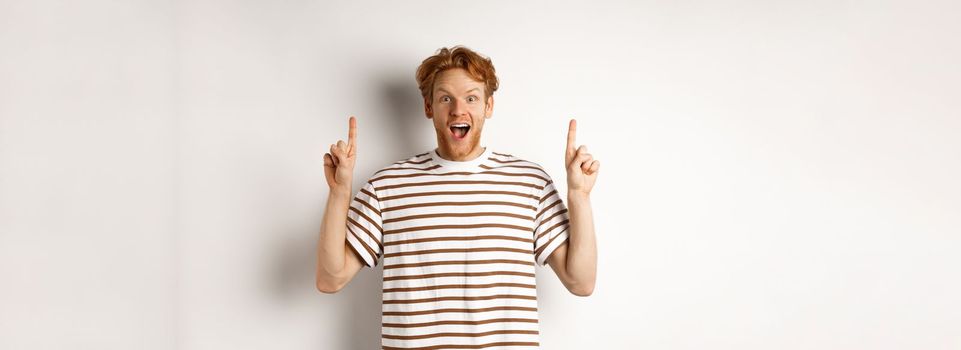 Amazed and excited redhead man checking out promotion, pointing fingers up and showing logo, staring at camera, standing over white background.