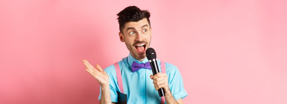 Image of male show host making speech, talking in microphone with clipboard under armpit, enteratin people on festive event, standing over pink background.